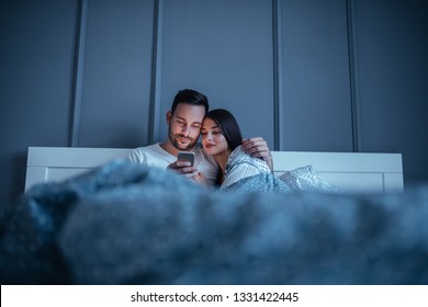 Happy young couple using a cellphone while lying in bed together at home - Powered by Shutterstock