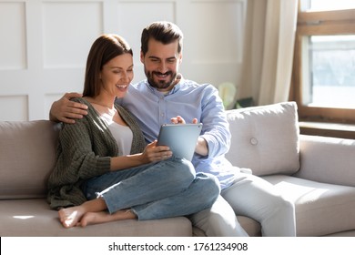 Happy Young Couple Using Computer Tablet Together, Sitting On Cozy Couch At Home, Smiling Beautiful Woman And Man Looking At Screen, Surfing Internet, Chatting Online In Social Network