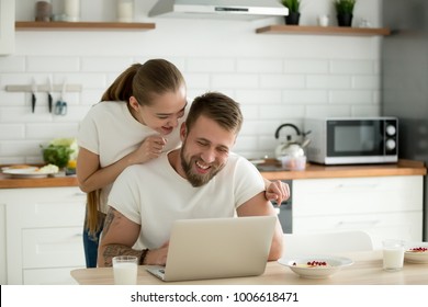 Happy Young Couple Using Laptop In The Kitchen Before Having Breakfast Together In The Morning, Smiling Man And Woman Looking At Computer Screen Checking Latest News Online In Social Networks