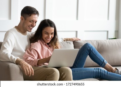 Happy Young Couple Using Laptop Together, Having Fun On Weekend, Sitting On Cozy Couch At Home, Laughing Man And Woman Looking At Computer Screen, Watching Funny Video Or Comedy Movie On Date