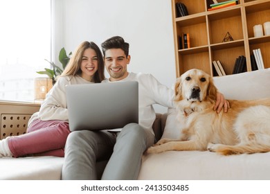 Happy young couple using laptop computer shopping online, watching video sitting with dog on comfortable sofa. Smiling man and woman working from home - Powered by Shutterstock