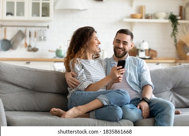 Happy young couple using smartphone together, relaxing on cozy couch at home, smiling overjoyed woman and man hugging, enjoying leisure time with gadget, sitting on sofa in living room - Powered by Shutterstock