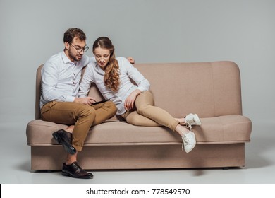 Happy Young Couple Using Smartphone Together While Sitting On Sofa Isolated On Grey 