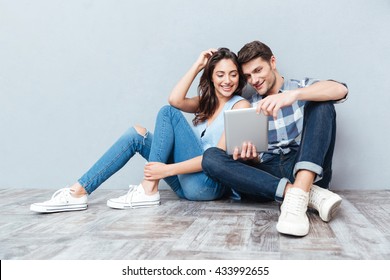 Happy young couple using tablet sitting on the floor at home isolated on gray background - Powered by Shutterstock