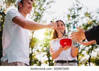 Happy Young Couple Volunteer In White Uniform Making Charity Mission Of Feeding Poor Homeless Man Giving Him Some Food And Bottle Of Water.