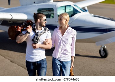 Happy Young Couple Walking From A Private Air Plane Walking On Runway With Brown Leather Luggage Bags. Models Wearing Casual Clothes And Smiling. Concept Is Travel On Vacation