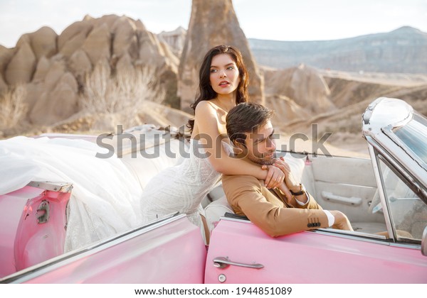 Happy young couple in wedding outfits\
sitting in pink retro car with background of rock formations at\
Cappadocia region. True love of young\
people.