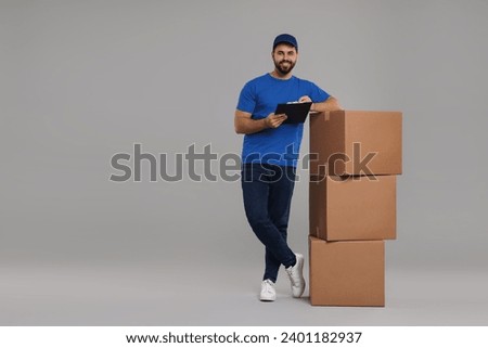 Happy young courier with stack of parcels and clipboard on grey background, space for text
