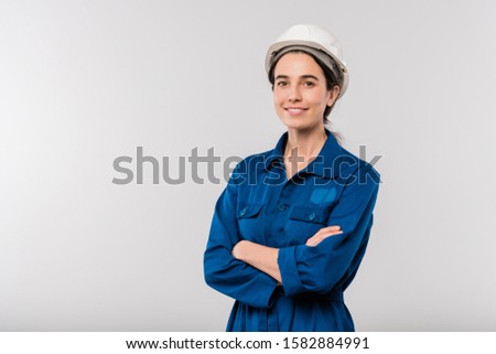 Happy young cross-armed female engineer in blue workwear and hardhat standing in isolation
