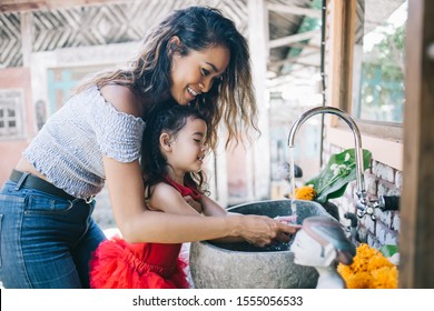 Happy Young Curly Asian Woman With Little Girl Washing Hands In Sink Together On Outside Terrace In Ethnic Style Smiling.  Protection And Hygiene, Enjoying Health Care 