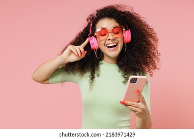 Happy young curly latin woman 20s wears mint t-shirt sunglasses eyes closed listen music in headphones hold in hand use mobile cell phone isolated on plain pastel light pink background studio portrait - Powered by Shutterstock
