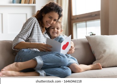 Happy young curly mother relaxing on sofa, embracing small preschool daughter, reading congratulations wishes in birthday card, feeling proud. Excited mommy enjoying greetings in postcard with child.