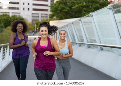 Happy young curvy women jogging together on city bridge. Healthy girls friends running on the city street to lose weight. Group of multiethnic oversize women running with building in the background.