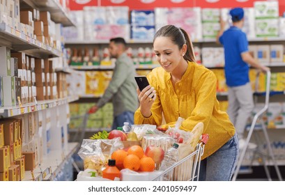 Happy young customer doing grocery shopping at the supermarket, she is leaning on a full shopping cart and using a smartphone - Powered by Shutterstock