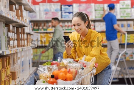Happy young customer doing grocery shopping at the supermarket, she is leaning on a full shopping cart and using a smartphone