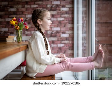 Happy young cute pretty girl with pigtails long hair sat at table indoors looking through the  with big happy smile and feet on window on bright summer day  during Covid 19 Coronavirus lockdown