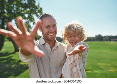 Happy Young Dad Holding His Little Son While Playing With Him In The Park On A Summer Day. Father And Kid Reaching Out Their Hands To Camera. Fatherhood, Childhood, Happiness Concept