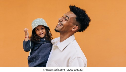 Happy Young Dad Laughing Joyfully While Carrying His Daughter In A Studio. Cheerful Single Father Celebrating Father's Day With His Adorable Daughter. Dad And Daughter Standing Against A Studio Backgr