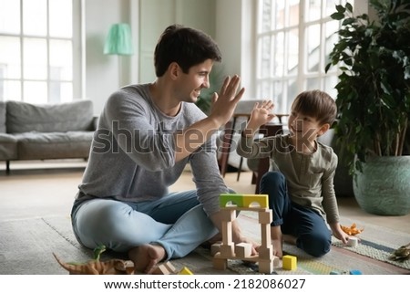 Happy young daddy giving high five to joyful small baby son, finishing constructing toy building with blocks. Bonding loving two male generations family involved in creative domestic activity.