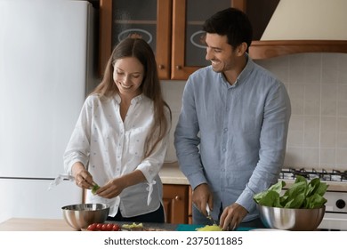 Happy young dating couple cooking salad for dinner together, chopping fresh vegetables into metal bowl on kitchen table, preparing natural ingredients, organic food, keeping healthy lifestyle - Powered by Shutterstock