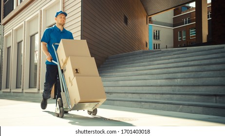 Happy Young Delivery Man Pushes Hand Truck Trolley Full of Cardboard Boxes and Packages For Delivery. Professional Courier Working Efficiently and Quickly. In the Background Stylish Modern Urban Area