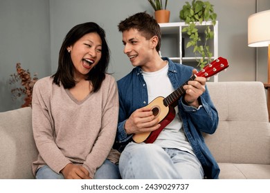 Happy young diverse couple singing a song playing guitar on sofa in living room at home. Lovely young adult woman and man having fun with ukulele in their apartment. - Powered by Shutterstock