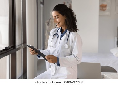 Happy young doctor woman reading medical records, getting report, optimistic test results for patient, reading paper document, looking at pad, smiling, leaning at workplace table in clinic office - Powered by Shutterstock