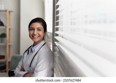 Happy young dreamy successful Indian female general practitioner doctor nurse medical worker looking in distance out of window, visualizing career opportunities or enjoying break time in clinic