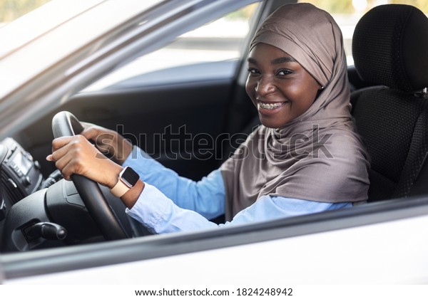 Happy Young Driver. Smiling black muslim lady in\
headscarf posing in her new car, got driving license, holding\
steering wheel and smiling, looking at camera through window,\
closeup shot, side view