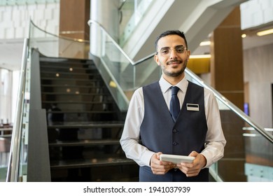 Happy Young Elegant Worker Of Contemporary Hotel Using Tablet While Standing Against Staircase And Looking At You Inside Lounge