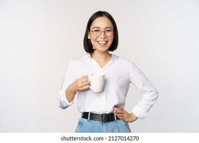 Happy young energetic asian woman smiling, drinking, holding cup mug of coffee, standing confident against white background - Shutterstock ID 2127014270