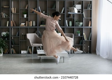 Happy young energetic beautiful Indian woman in ballet skirt making elegant moves, dancing to classic music in modern living room, enjoying domestic hobby activity alone on weekend.