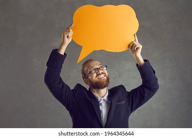 Happy Young Entrepreneur Or Office Worker In Suit Holding Up Orange Thought Bubble Isolated On Grey Background. Smiling Guy Shows Mockup Paper Speech Balloon, Sharing Message And Expressing Opinion