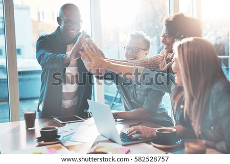 Happy young entrepreneurs in casual clothes at cafe table or in business office giving high fives to each other as if celebrating success or starting new project