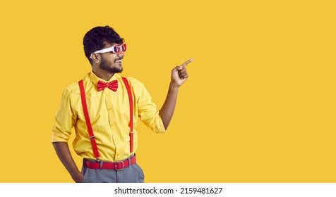 Happy young ethnic guy shows something on yellow copy space background. Cheerful Indian man in shirt, glasses, bowtie and pants with suspenders smiles and points his index finger to copyspace side - Shutterstock ID 2159481627