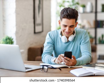 Happy Young Ethnic Man With Headphones Smiling And Browsing Social Media On Smartphone During Break In Work At Home
