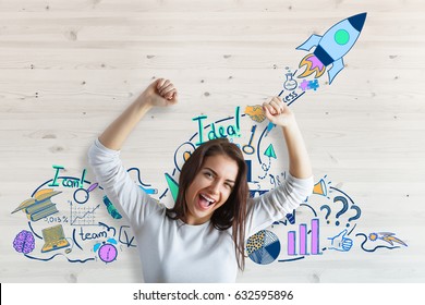 Happy Young European Businesswoman Celebrating Success On Wooden Background With Rocket Sketch. Entrepreneurship Concept