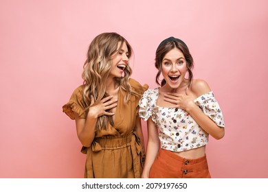 Happy young fair-skinned girls laugh while enthusiastically holding their breasts with their hand. Standing on pink background, teenagers with curly hair are dressed in light summer clothes.
