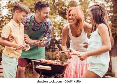 Happy Young Family Barbecuing Meat On The Grill.