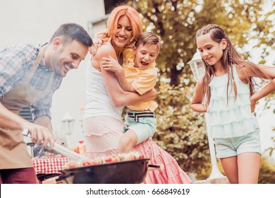 Happy Young Family Barbecuing Meat On The Grill.