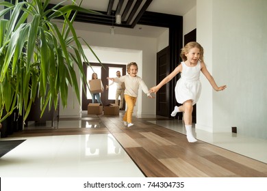 Happy Young Family With Cardboard Boxes In New Home At Moving Day Concept, Excited Children Running Into Big Modern Own House Hallway, Parents With Belongings At Background, Mortgage Loan, Relocation 