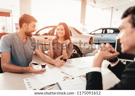 Happy Young Family Are Choosing A New Car In Showroom. Dialogue With Dealer. Cheerful Customer. Automobile Salon. Make A Decision. Cup Of Coffee. End Of A Deal. Good Offer. Buyer And Seller.