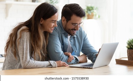 Happy Young Family Couple Bonding Laughing Using Laptop Looking At Screen Sit At Table, Smiling Husband And Wife Buying Choosing Goods Online Doing Internet Shopping Order With Delivery At Home