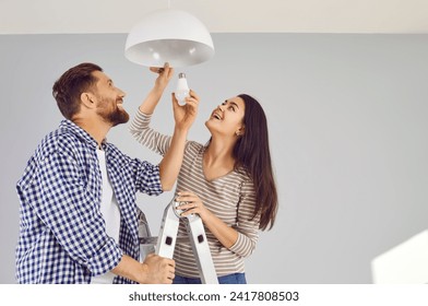 Happy young family couple changing a light bulb together. Cheerful, smiling man and woman standing on a step ladder at home and placing a modern energy saving lightbulb in a white lamp on the ceiling - Powered by Shutterstock