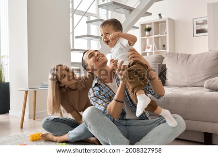 Happy young family couple having fun playing with cute small toddler kid son in modern living room together. Smiling parents mum and dad enjoying spending time with cute funny child boy at home.