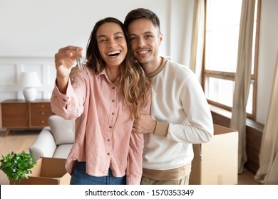 Happy young family couple holding key to new home on moving day concept, first time real estate owners man husband embrace woman wife look at camera proud buying property stand in own flat with boxes - Shutterstock ID 1570353349