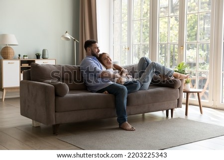 Happy young family couple relaxing on comfortable couch together, hugging, talking, laughing, enjoying leisure, being in new cozy home interior with terrace, big window