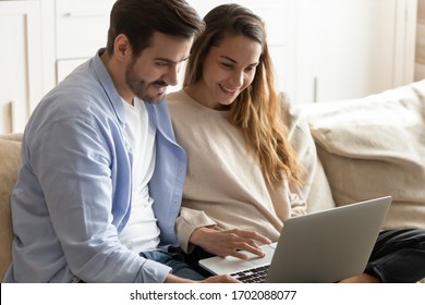 Happy young family couple sitting on couch, using computer together at home. Smiling spouses shopping online in internet store, web surfing information or chatting with friends in social networks. - Powered by Shutterstock