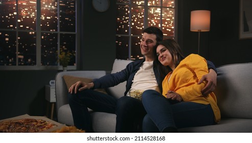 Happy Young Family Couple Watching Tv At Home, Sitting On Couch Eating Popcorn. Loving Man And Woman Resting On Sofa Enjoying Movie Night.