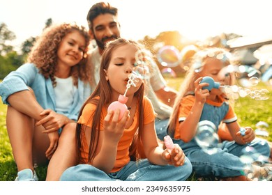 Happy young family dad, mom and two daughters blowing soap bubbles sitting in the meadow on the grass in the park, playing and having fun together on a warm sunny day off.